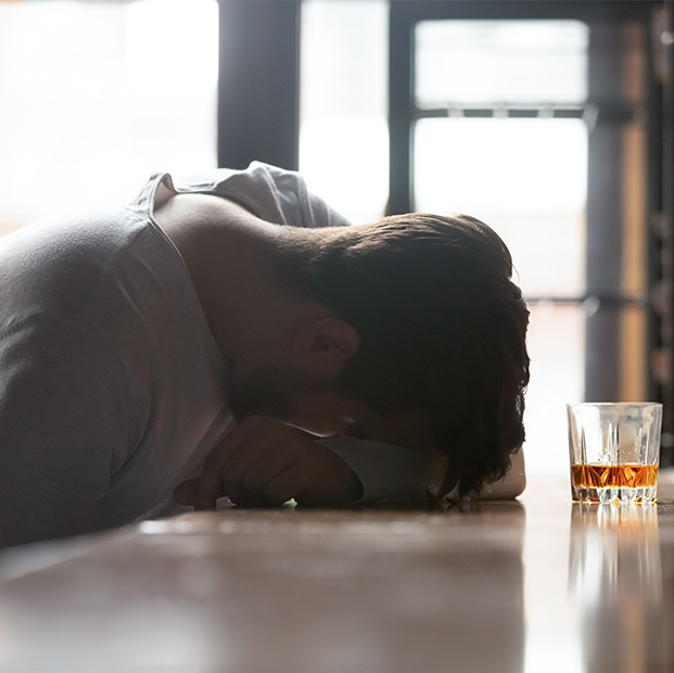 A man sits at a bar with his head down, next to a glass of alcohol. This image represents someone who may be in need of substance use treatment online in San Francisco and Los Angeles offered by Dr. Nathan Brandon.