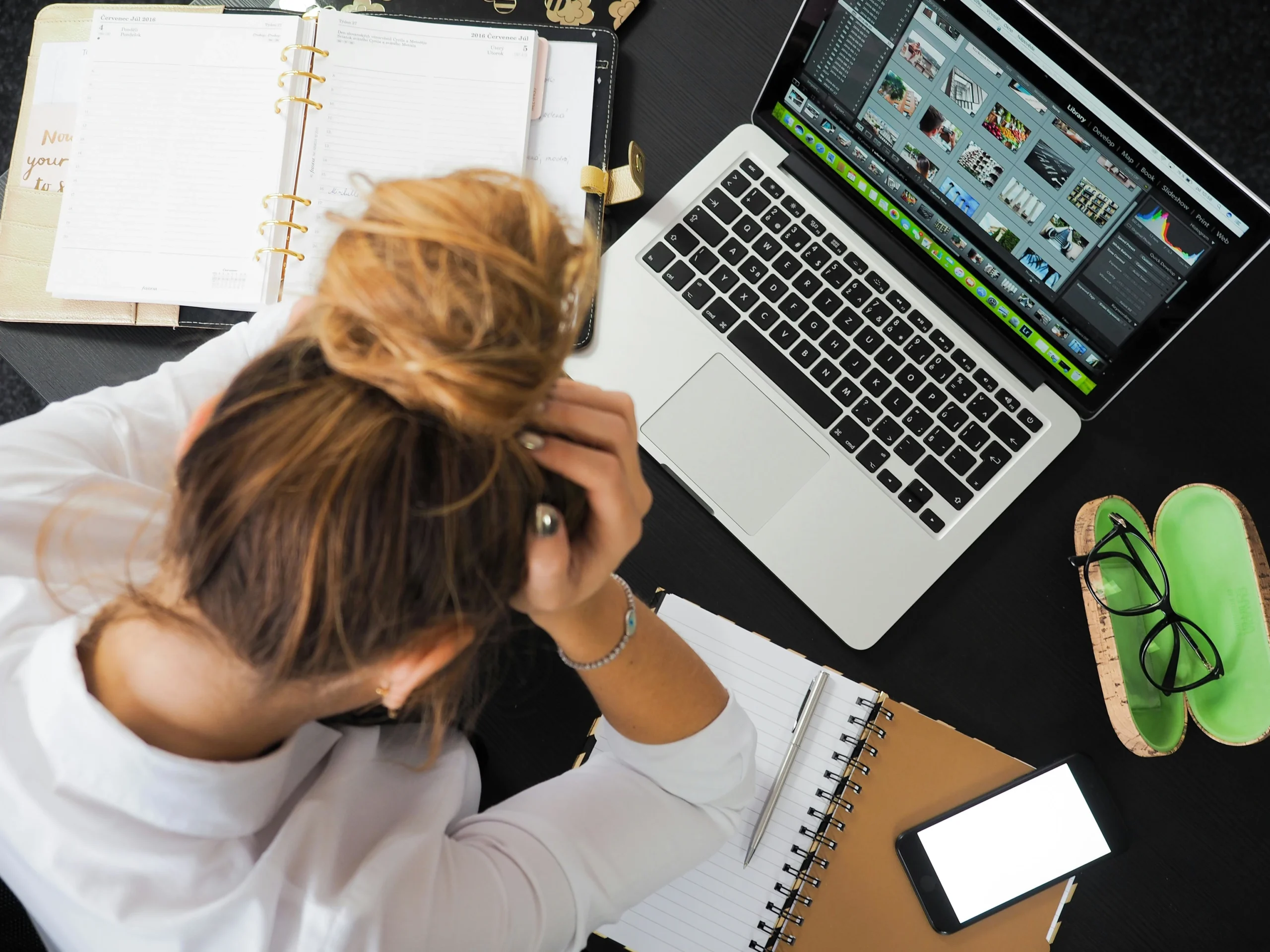 A woman with a messy bun sits at a desk, stressed, surrounded by a laptop, notebooks, a phone, and glasses.
