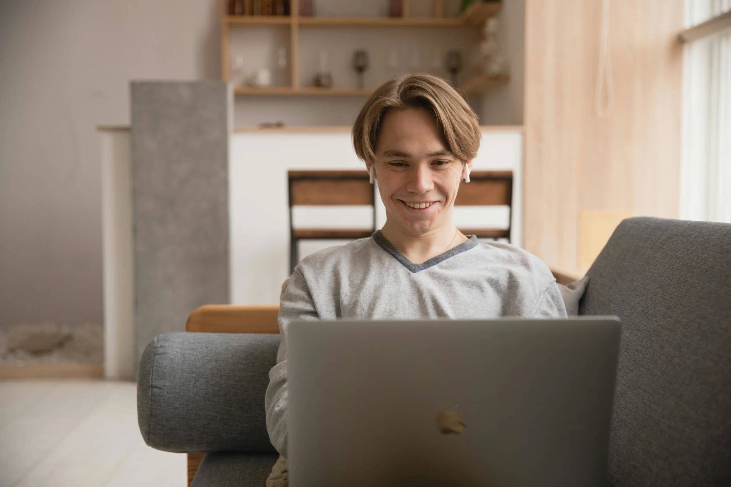 A person in a gray sweater sits on a sofa, focused on a laptop in a modern, cozy living space with soft lighting.